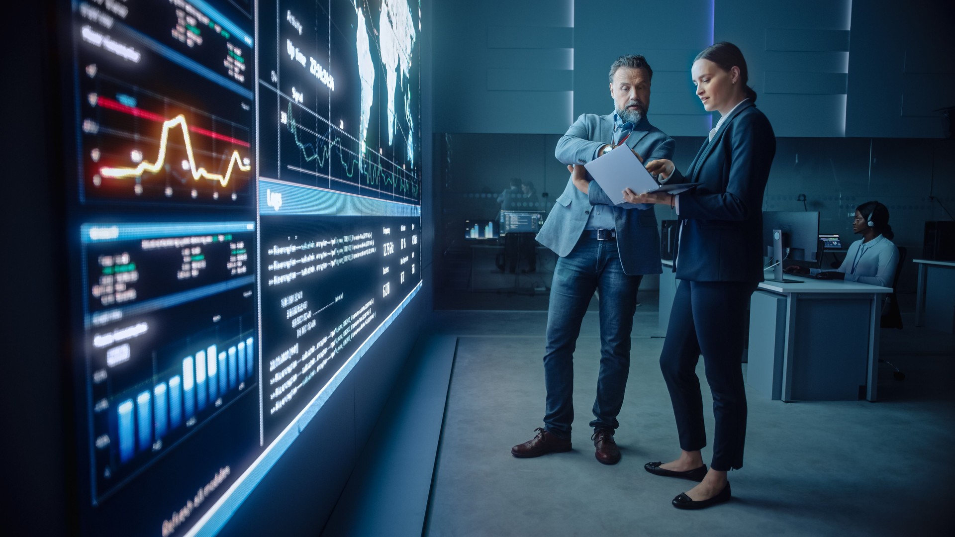 El gerente de proyecto y el ingeniero de ciencias de la computación hablan mientras usan la pantalla grande y una computadora portátil, mostrando datos de infografías de infraestructura. Sala de Control y Monitoreo de Sistemas de la Empresa de Telecomun